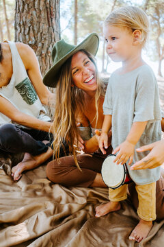 Delighted mother and crop father teaching little kid playing bongo drum while spending time together in woods