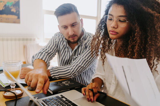High angle of content multiracial couple of entrepreneurs sitting at table and reading paper documents while working from home