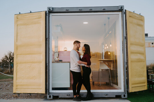 Side view of smiling couple cuddling near window of modern container house at sunset in countryside