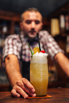 Soft focus of male mixologist putting glass of cold exotic cocktail with fruits and ice on counter during work in bar looking at camera