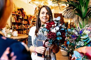 Professional female florist holding flower bouquet while helping cropped unrecognizable costumer in decor shop looking at each other