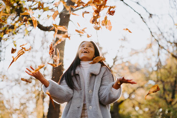 Low angle of carefree ethnic female in warm coat tossing fallen autumn leaves in park on sunny day while having fun and looking up