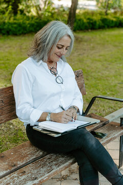 Middle aged female entrepreneur in smart casual wear sitting on bench and writing list of daily routine tasks while working remotely in park