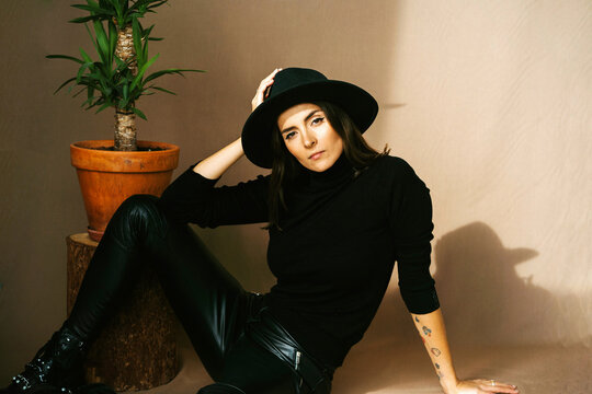 Confident young tattooed female in stylish black leather pants and turtleneck with cowboy hat sitting on floor near potted plant and looking at camera in light studio
