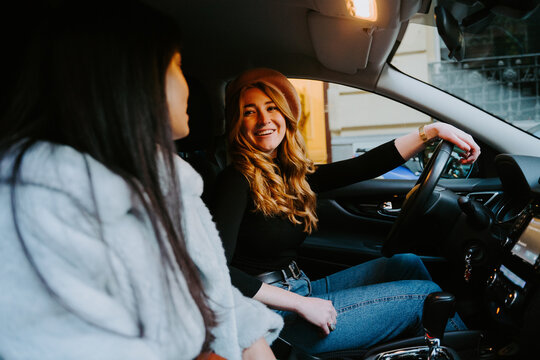 Cheerful female friends sitting on driver and passenger seat in contemporary car while talking and looking at each other