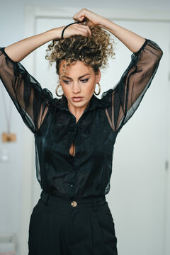 Serious female with curly hair and in elegant black clothes standing in room and making ponytail while looking away