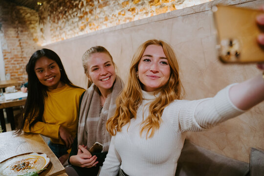 Group of cheerful diverse female friends sitting at table in cafe and taking selfie on smartphone while spending weekend together
