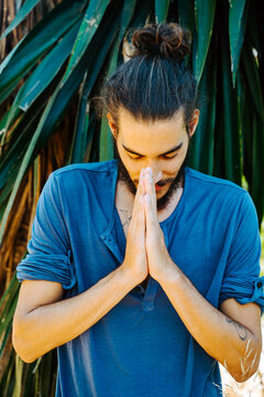 Grateful ethnic male with long hair in bun standing with praying hands with closed eyes and doing yoga in tropical park