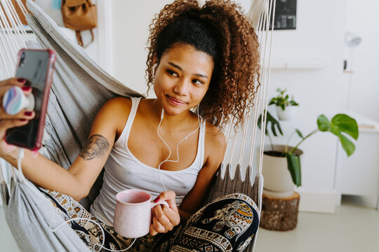 Smiling young African American curly haired female listening to music through earphones and taking selfie on smartphone while chilling in hammock at home