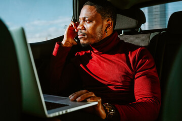 Busy African American businessman sitting in luxury car with laptop and speaking with client on smartphone while looking away