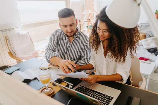 High angle of content multiracial couple of entrepreneurs sitting at table and reading paper documents while working from home