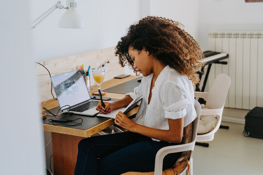 Side view of modern young African American female remote employee in casual outfit sitting at table with laptop and writing in planner while working in home office