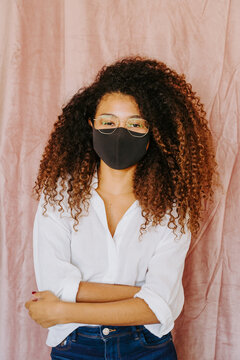 Young ethnic female with Afro hairstyle standing on pink background and putting on protective mask from coronavirus while looking at camera
