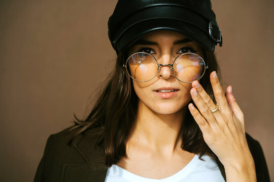 Modern young female model in stylish black leather cap and trendy round glasses looking at camera against brown background
