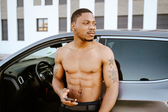 Fit African American male with six pack standing with smartphone near luxury automobile and looking away