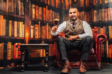 Full body of man in elegant suit holding glass of Irish coffee while resting in comfortable armchair looking at camera