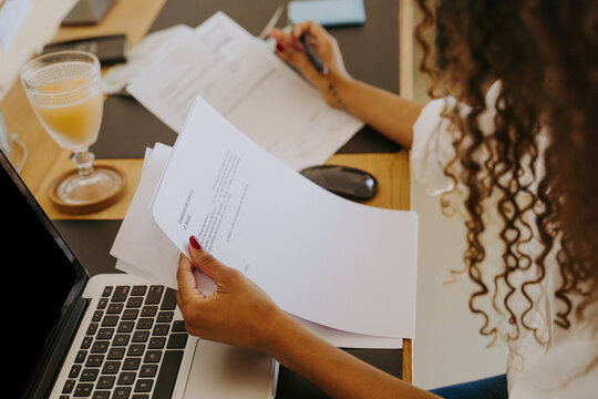 High angle side view of female bookkeeper sitting at table in home office and analyzing financial reports and paper documents during remote work