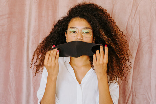 Young ethnic female with Afro hairstyle standing on pink background and putting on protective mask from coronavirus while looking at camera