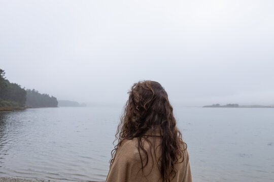 young woman from behind with a brown cardigan looking at a beautiful beach on a cloudy day
