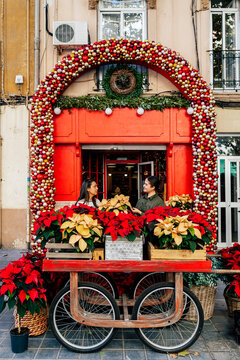 Woman standing outside shop with female costumer at red door with christmas decorative baubles holding pot of poinsettia flower in daylight