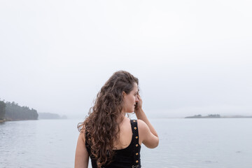 Portrait of a white woman in her 30s.
Has curly hair.
He is wearing a black jumpsuit.
You can see the beach in the background.
It is the coast of Galicia, Spain