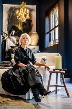 Full body of stylish middle aged lady in elegant black dress and high heeled boots sitting on chair in living room with vintage interior design