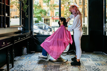 Side view of female hairdresser using curling iron and making trendy hairstyle for client in modern salon during coronavirus pandemic