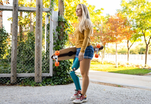 Positive female skater with bionic leg prosthesis walking with longboard in city and looking away