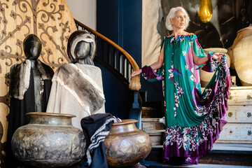Confident mature female in elegant maxi dress standing near stairs in expensive house while looking at camera