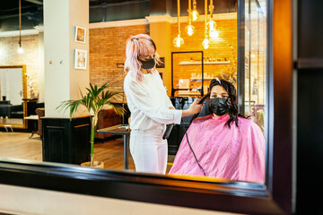 Side view of female hairdresser using curling iron and making trendy hairstyle for client in modern salon during coronavirus pandemic
