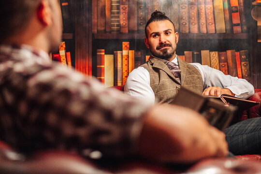 Male friends sitting in armchairs and choosing drink from menu while talking to each other in restaurant