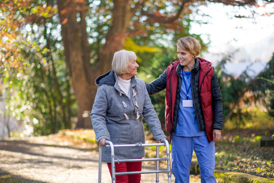 Senior woman with walking frame and caregiver outdoors on a walk in park.