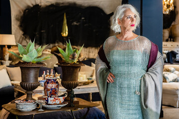 Confident mature female wearing sparkling glamour dress standing with hands on waist in luxury room in house and looking away