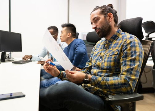 Side view of focused bearded male analyst manager in casual outfit examining paper with diagrams while working in modern workspace with colleagues