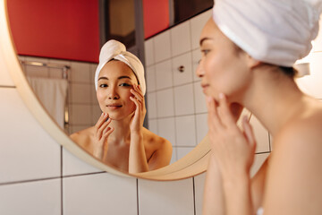 Close-up portrait of girl in towel on her head looking into mirror. Woman without make-up gently touches face