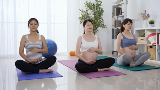 three of healthy and fitness asian pregnant women seated on exercise mat are taking group yoga lessons.