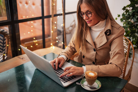 Smiling young female blogger in casual clothes and glasses sitting at table with cup of coffee and typing on laptop keyboard while preparing content for social networks in cozy cafeteria