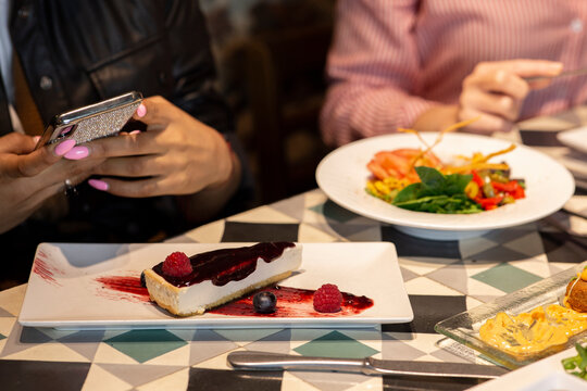 Crop anonymous female taking picture of piece of appetizing cheesecake with berries on mobile  phone while having dinner with friends in restaurant