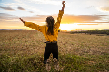Back view os unrecognizable carefree female in casual outfit in moment of jumping above ground in meadow on background of sunset in rural area