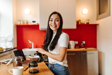 Brown-eyed girl in white T-shirt smiles, looks into camera and makes coffee