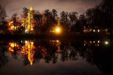 Fototapeta premium A historic village church that is illuminated by lamps in Berlin-Lichtenrade at night and that is reflected in a pond.
