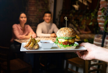 Crop anonymous waiter holding tray with delicious appetizing burger served with baked potatoes and sauce while serving meal for clients in cafe