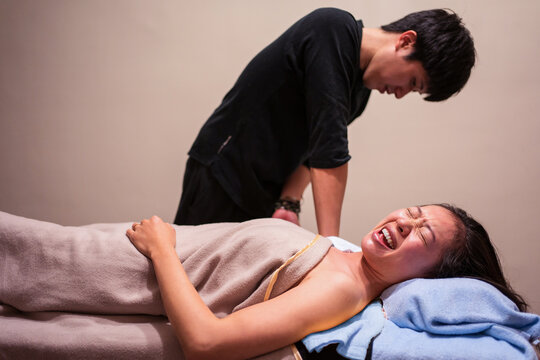 Side view of young Asian female patient crying in pain while getting professional osteopathic massage from therapist in clinic