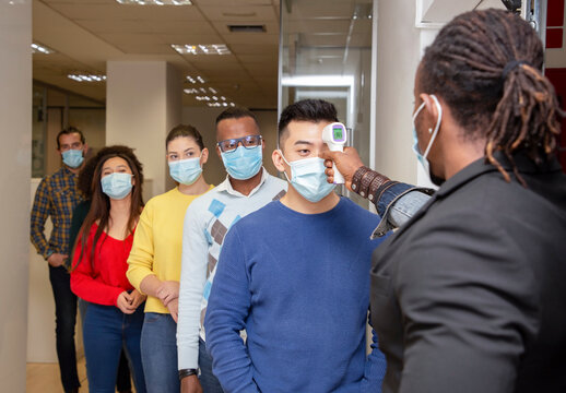 Group of multiracial colleagues in casual clothes and medical masks passing through procedure of body temperature control for coronavirus prevention before entering workplace