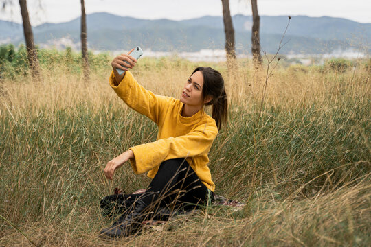 Side view of charming female in yellow sweater sitting in dry field and taking selfie while entertaining at weekend in countryside