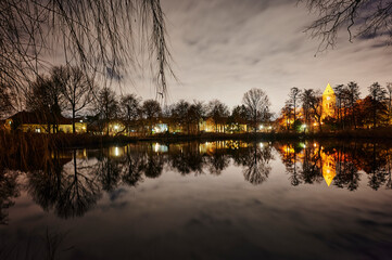 A historic village church that is illuminated by lamps in Berlin-Lichtenrade at night and that is reflected in a pond.