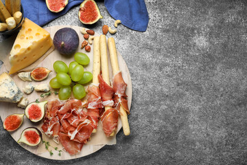 Board with delicious figs, cheese, grapes and bread with proscuitto on grey table, flat lay. Space...