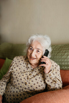 Smiling Senior Female Sitting On Comfortable Couch And Speaking On Mobile Phone With Friend While Looking Away