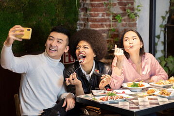 Group of positive young multiracial friends having fun and taking selfie on smartphone while enjoying delicious meal together in cozy cafe