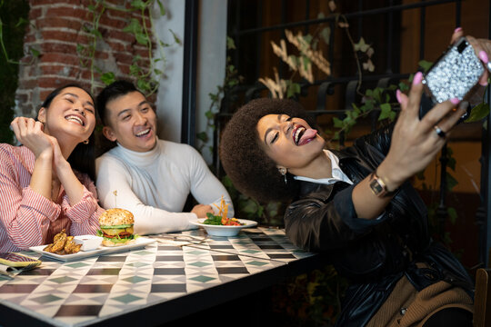 Cheerful young African American female showing tongue while taking selfie on smartphone with laughing fiends during dinner in cozy restaurant